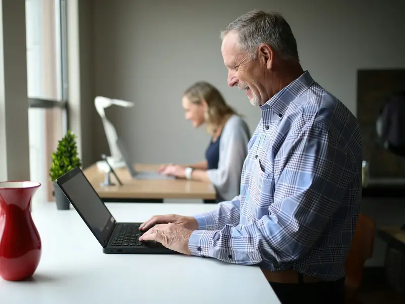 An older man smiling as he works on a laptop in an office, with a colleague at a desk behind him.