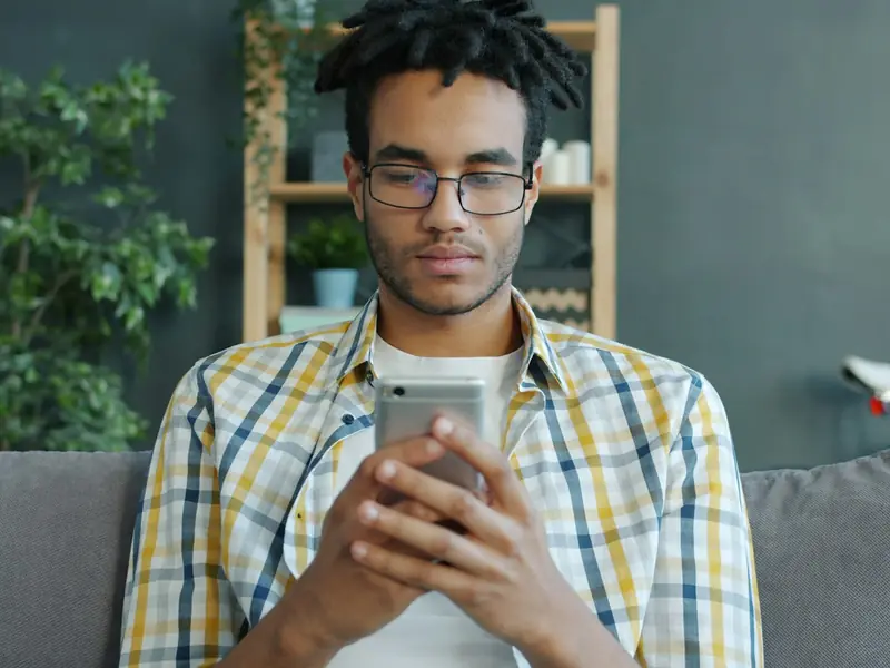 A young man with glasses sitting on a sofa at home, reading something on his phone.